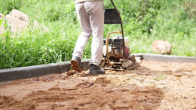Person beside plate compactor on sandy construction ground near fresh concrete border, with dirt mound, green vegetation, rough soil texture, and bright daylight, showing site