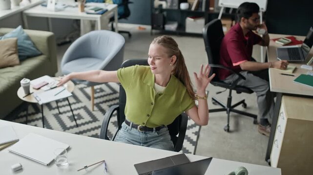 Overhread medium shot of young Caucasian woman stretching arms and dancing in office chair while taking break from work, slow motion. Ideal for corporate wellness and employee satisfaction campaigns