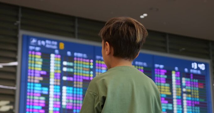 Young passenger standing with back to camera in front of large digital display at airport terminal, intently checking arrival flights. Selective focus on boy with timetable blurred on background