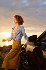 Fototapeta premium Young woman with curly hair strolls beside a sleek car at sunset, enjoying a warm breeze, golden light, casual denim jacket, striped top, confident relaxed mood.