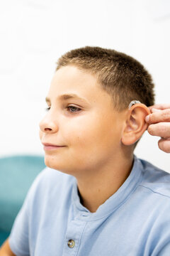 Vertical photo of a child wearing a behind-the-ear hearing aid while a professional adjusts the device in a clinical setting. Concept of hearing health, medical technology, hearing loss.