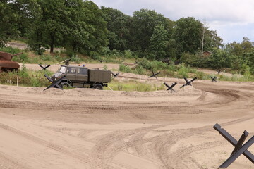 A preserved military truck displayed on a sandy training ground with defensive barriers. The vehicle is part of the outdoor military museum exhibition.  © Fullip