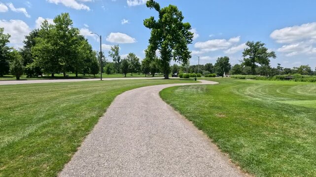 POV Walking on Recreational Public Park Trail. A first-person view moving down a curved pedestrian walking path lined by beautiful manicured lawns and mature trees in Forest Park, St. Louis, Missouri.