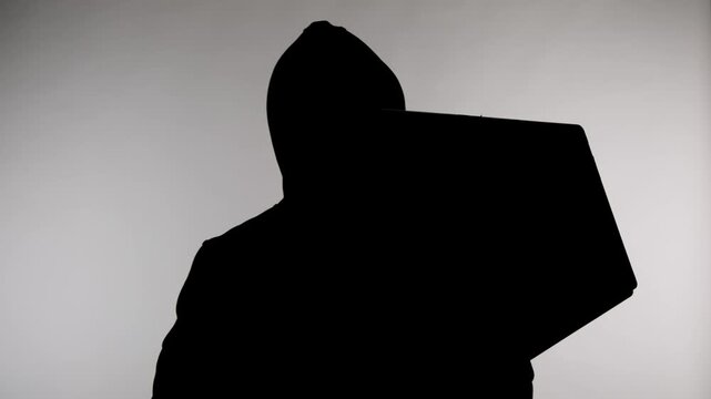 An anonymous silhouette of a man standing on a white background wearing a hoodie and working on a laptop