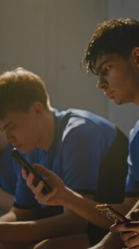 Boys in Sports Uniforms Sitting on Locker Room Bench. Teenager Absorbed in Own Smartphone, Scrolling Through Social Media or Apps Before Training. Technology Addiction, Screen Time. Vertical Shot.