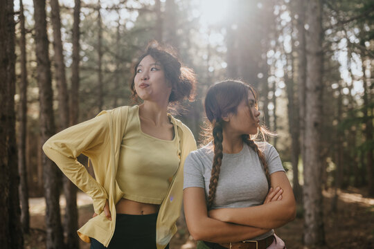 Two young women stand in a sunlit forest, one with hands on hips and the other with arms crossed. The pair look in opposite directions, captured in a candid outdoor moment with backlit trees.