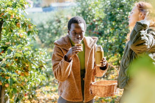 Happy diverse friends drinking tea from thermos in sunny autumn apple orchard
