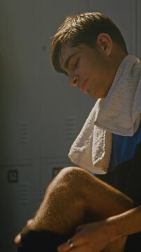 Teenage Player in Uniform Sits on Bench in Dressing Room, Putting on Black Neoprene Knee Brace. Sports Safety, Injury Prevention, and Athletic Preparation Before Training or Match. Vertical Shot.
