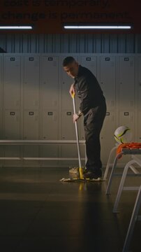 Janitor in Uniform Meticulously Cleans Locker Room After Sports Match. Technician Methodically Moves Flat Mop Across Tiled Floor, Ensuring Cleaning and Proper Sanitation Under Benches. Vertical Shot.