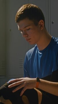 Teenage Sitting in Locker Room With Knee Meticulously Wrapped in Athletic Kinesiology Tape. Boy Looks Down Injured Joints, Fingers Carefully Adjusting Black Neoprene Knee Brace Sleeve. Vertical Shot.