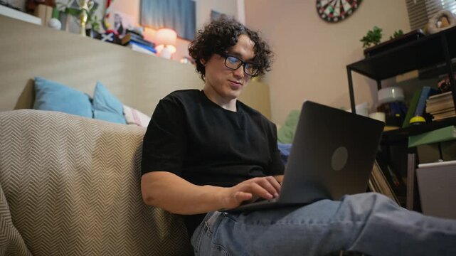 Young adult Asian man sitting on floor, typing on laptop computer in home office. Footage is ideal for commercials promoting remote work, online education, or freelance lifestyle, low angle