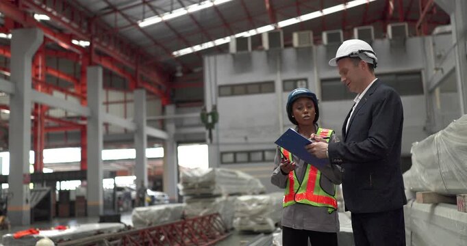A senior manager and an African female worker in a safety vest use a digital tablet to inspect steel coils stored in a warehouse under an overhead crane hook discussing stock quality