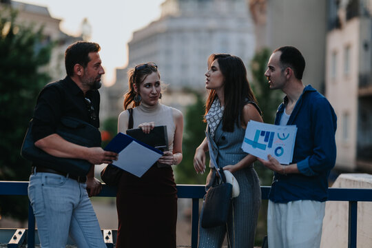 Four business colleagues review reports and charts while conversing on a city bridge. A small outdoor meeting shows teamwork and discussion with papers and a tablet.