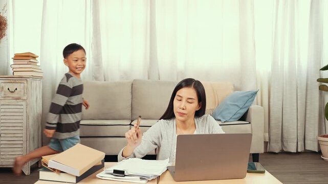 Asian mother experiences extreme stress and exhaustion while trying to work from home on laptop computer. Young girl and playful boy running around, causing a loud distraction in the living room