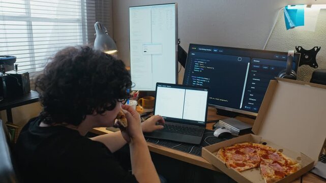 Asian young adult man coding on laptop and eating pizza at home office desk with multiple monitors. Clip represents software development, remote work lifestyle, and IT education concepts, handheld