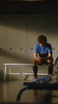 Sad Teenager Sits Isolated From Teammates. Supportive Coach Approaches For Private Conversation. Realistic Atmosphere of Sports Team, Interaction in Background and Individual Struggle. Vertical Shot.