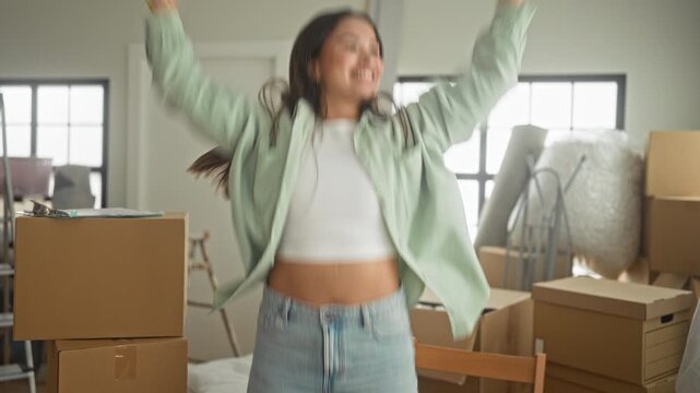 Woman raising arms among stacked moving boxes revealing bare midriff in building during unpacking and celebration; fresh start joy.