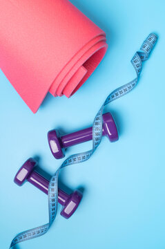 Pink fitness mat with purple dumbbells and measuring tape arranged on blue background with copy space. Fitness supports home workouts, wellness promotion, sports branding and healthy lifestyle 