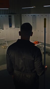Male Cleaner in Black Overalls Enters Empty Locker Room to Tidy Up After Athletic Team Leaves. Janitor Mops Tiled Floor With Mop, Checks Open Lockers. Facilities Maintenance Services. Vertical Shot.