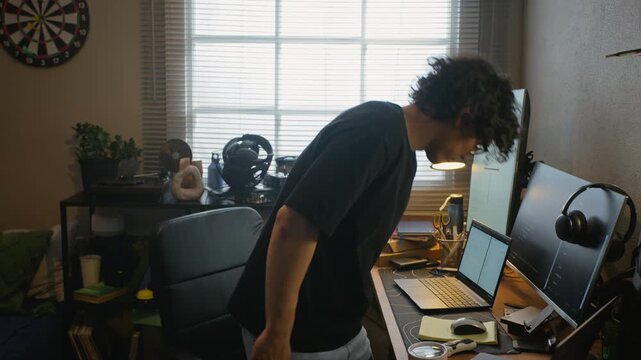 Young adult Asian man walking to desk, sitting down, and typing on laptop in home office. Footage represents remote software development, freelance programming, or online coding bootcamps