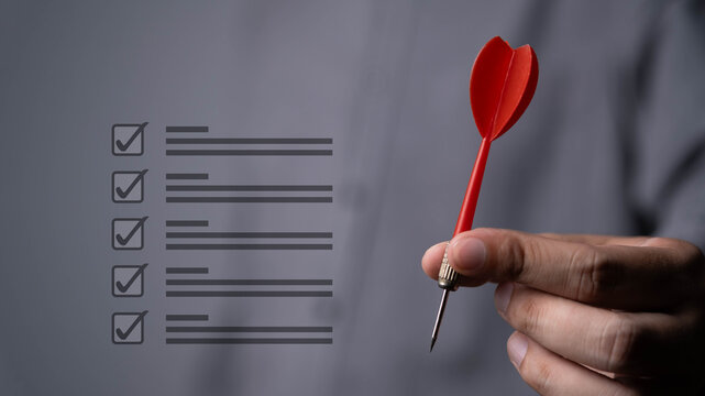 Close-up of a hand holding a red dart next to a checklist interface, symbolizing goal achievement, business planning, task management, strategic focus, project execution, and performance improvement.