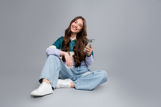 Smiling young woman sitting on floor with smartphone in casual jeans and sneakers, relaxed lifestyle portrait showing social media and communication