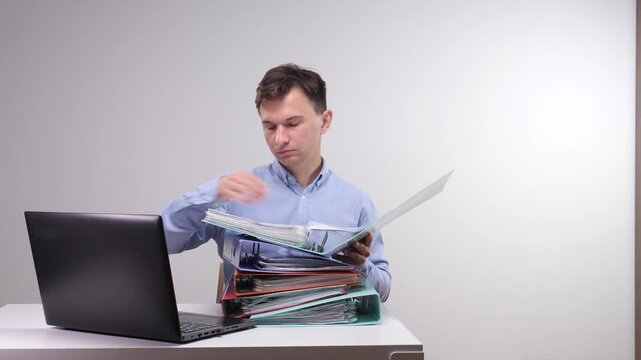 Man in a blue shirt managing a large pile of document binders while working at his laptop, focusing on detailed administrative tasks