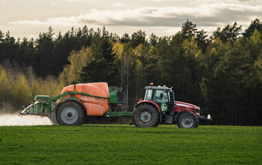 Obraz premium Tractor With Mounted Sprayer Applying Fertilizer On Green Field Near Forest At Dusk