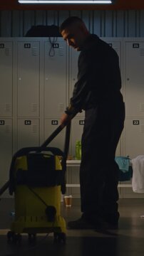 Janitor Cleans Up Heavily Dressing Room After Football Match. Worker Uses Powerful Vacuum Cleaner Clear Dust, Debris From Floor, Where Players Left Discarded Towels, Clothes, Trash. Vertical Shot.