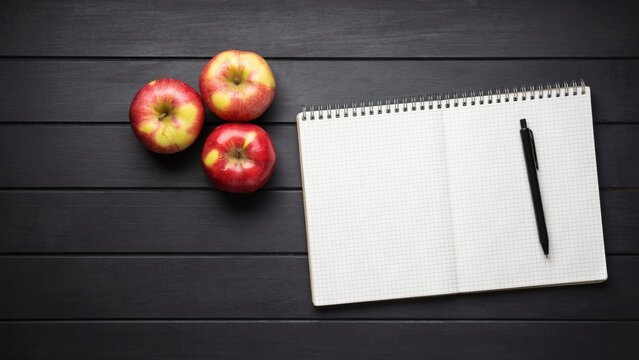 Three red apples and a spiral notebook with a pen on a black wooden table