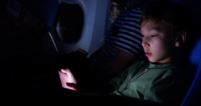 Young boy immersed in digital entertainment on tablet, interacting with it during nighttime airplane journey. He seated in same row with parents in dimly lit economy class