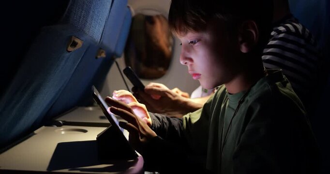 Young boy playing with music app on tablet placed on folding tray table during nighttime airplane journey, selective focus on boy. Dimly lit economy class cabin and parents in background