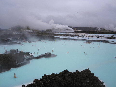 People enjoying the milky blue waters of the Blue Lagoon geothermal spa, surrounded by volcanic rocks and steaming industrial structures under a cloudy sky in Iceland