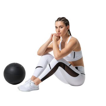 Athletic young caucasian woman with dutch braids wearing white patterned leggings seated by a medicine ball in a bright studio calm determination atmosphere