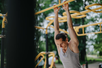 Fototapeta premium Young man in a gray tank top hangs from yellow monkey bars at an outdoor gym. He performs a calisthenics exercise focused on upper-body strength in a park setting.