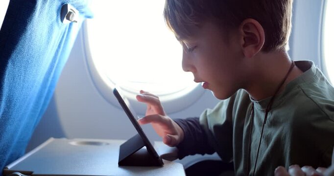 Young passenger sitting by airplane window, using digital tablet placed on foldable tray table, boy swipes his finger on touchscreen to move virtual object. Slow motion side view shot