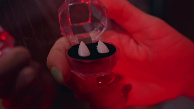 Close Up View of Artificial Vampire Fangs Displayed in Small Decorative Case Under Red Light, Halloween Costume Preparation, Horror Theme, Spooky Celebration