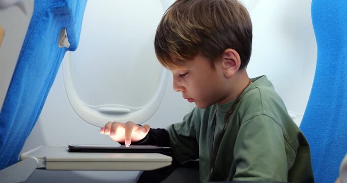Side view of young boy deeply engaged with learning app on tablet during airplane flight, showing range of emotions as he interacts with touchscreen. He tapping, pausing thoughtfully