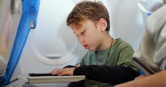 Young passenger boy engaging with tablet during flight, focused on solving something within learning app or educational game. When his mother points at screen to help, he gently resists her assistance