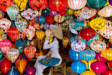 An Asian woman in an ao dai is sitting and admiring the colorful lanterns in the city of Hoi An, Vietnam. © chanchai