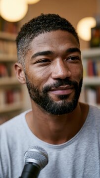 Man with short black hair and beard holding a microphone, wearing a gray t-shirt, seated in a library with bookshelves and soft lighting in the background