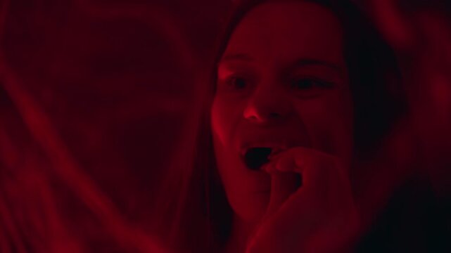 Young Woman Adjusting Artificial Vampire Fangs and Combing Hair in Front of Mirror Under Red Light During Halloween Costume Preparation, Spooky Transformation, Holiday Celebration