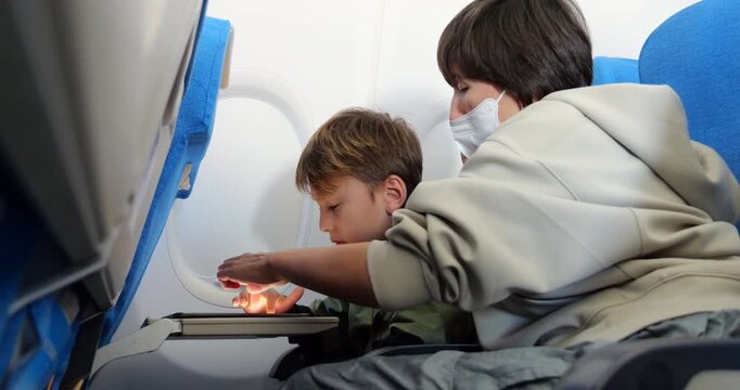 Young boy engaging with learning app or educational game on tablet during airplane flight, supported by his mother sitting beside him wearing protective surgical mask. Moment of in-flight time.