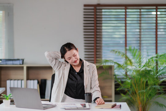 Young Asian businesswoman sitting at her office desk, feeling tired and stressed while stretching her neck to alleviate discomfort, pain, and tension from prolonged work