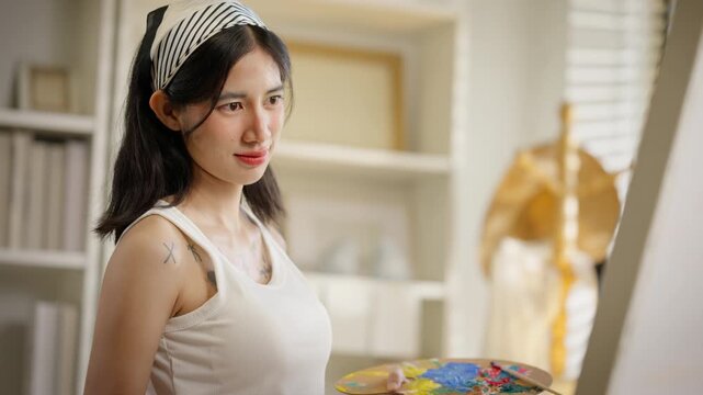 Close up of a beautiful young Asian woman painter smiling and focusing on her artwork. Talented female artist painting on canvas in home studio. Art therapy, inspiration, and mindfulness concept.