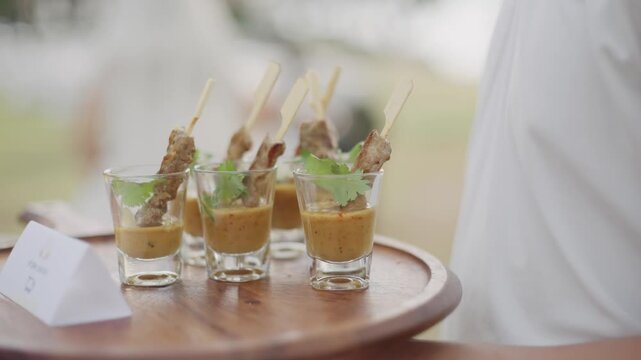A server carrying a wooden tray with delicious meat skewers in small glass cups, perfect for catering, cocktail parties, and luxury hospitality events.