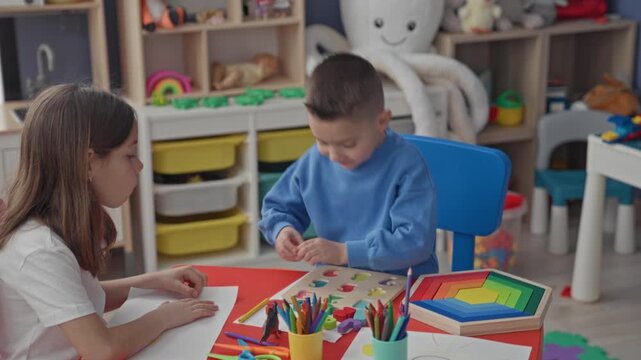 Girl and boy placing wooden puzzle pieces and drawing on paper at a small red table in a classroom building with crayons, colored pencils and a rainbow stacker visible; concentration playful learning.