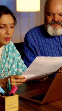 Asian Indian senior couple in their modern home during the evening, reviewing documents, bills, and papers while the husband assists his wife in updating information on the laptop