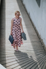 Woman wearing a pink polka dot dress and sunglasses walks up a sunlit textured ramp carrying a bag. The fashionable retro outfit and strong shadow create a chic urban street scene. © qunica.com