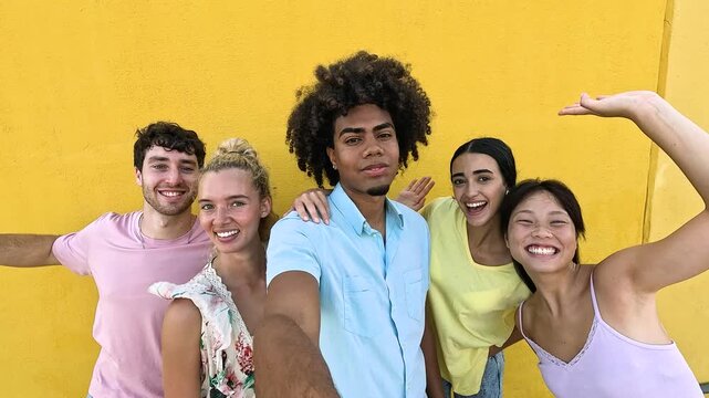 Vibrant, multiethnic group of young friends laughing and posing together, taking a selfie video against a bright yellow background